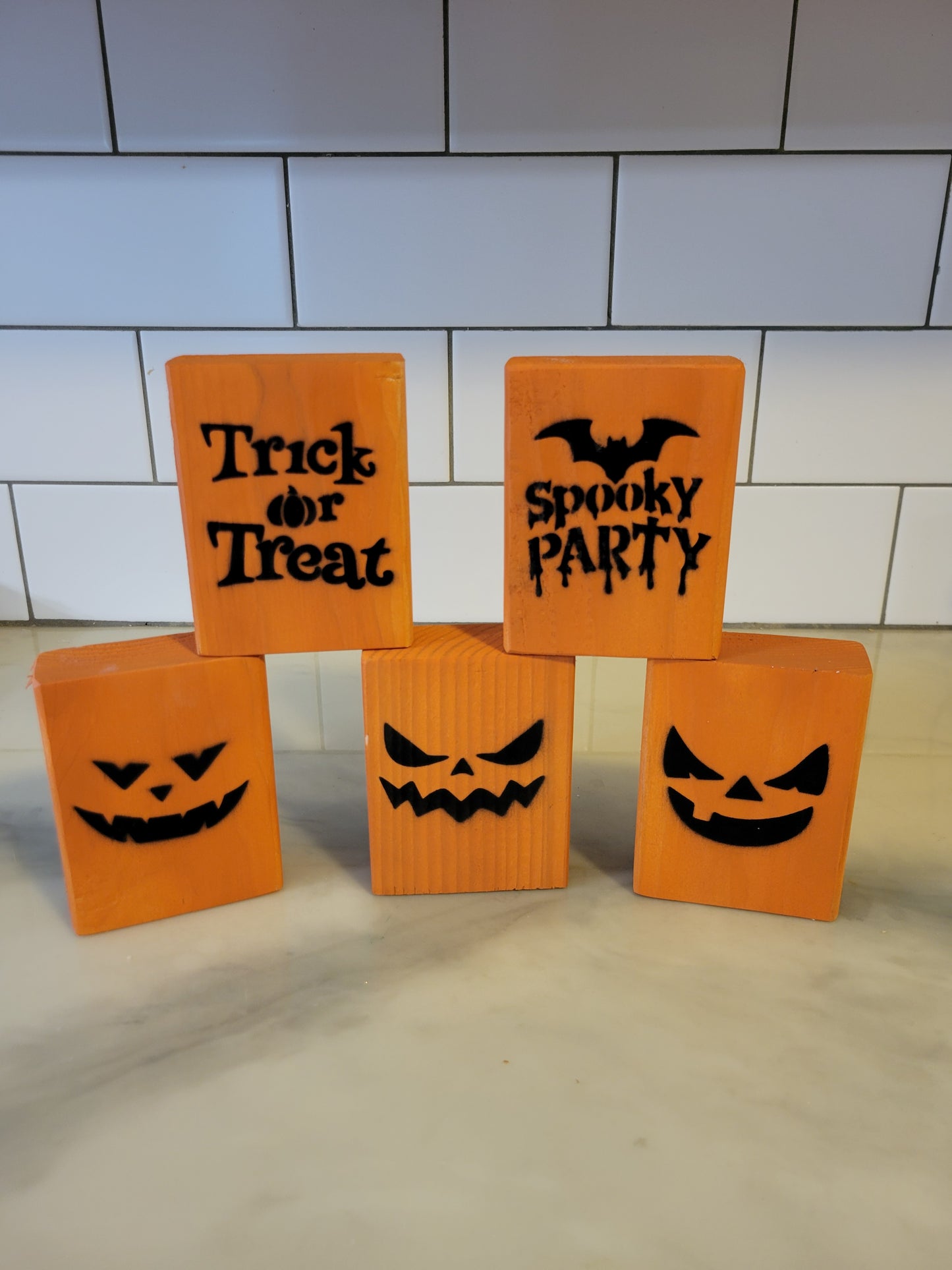 Five orange Halloween-themed decorative blocks arranged on a light countertop in front of a white subway tile backsplash. The blocks include jack-o'-lantern faces and text reading 'Trick or Treat' and 'Spooky Party'.