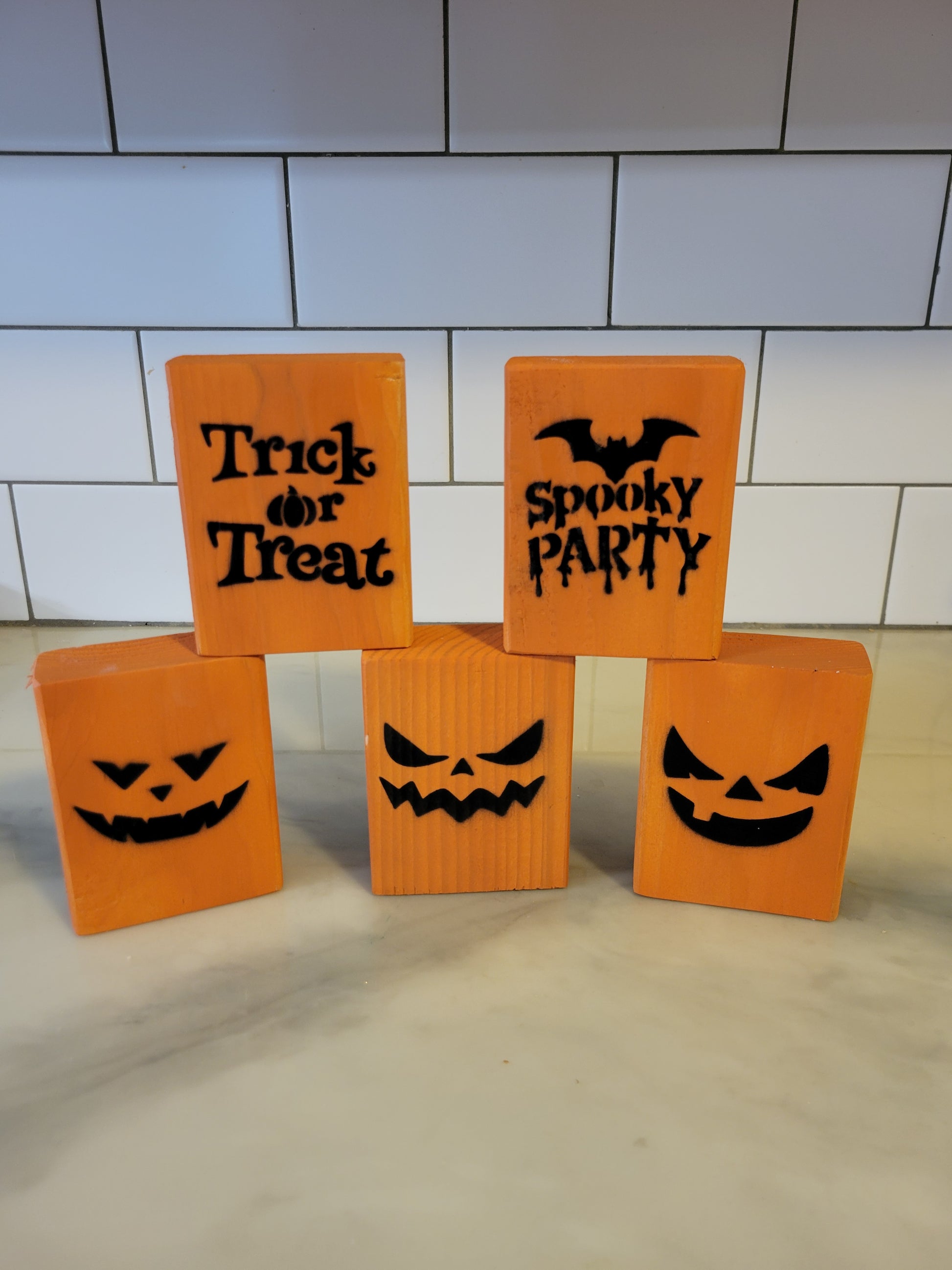 Five orange Halloween-themed decorative blocks arranged on a light countertop in front of a white subway tile backsplash. The blocks include jack-o'-lantern faces and text reading 'Trick or Treat' and 'Spooky Party'.