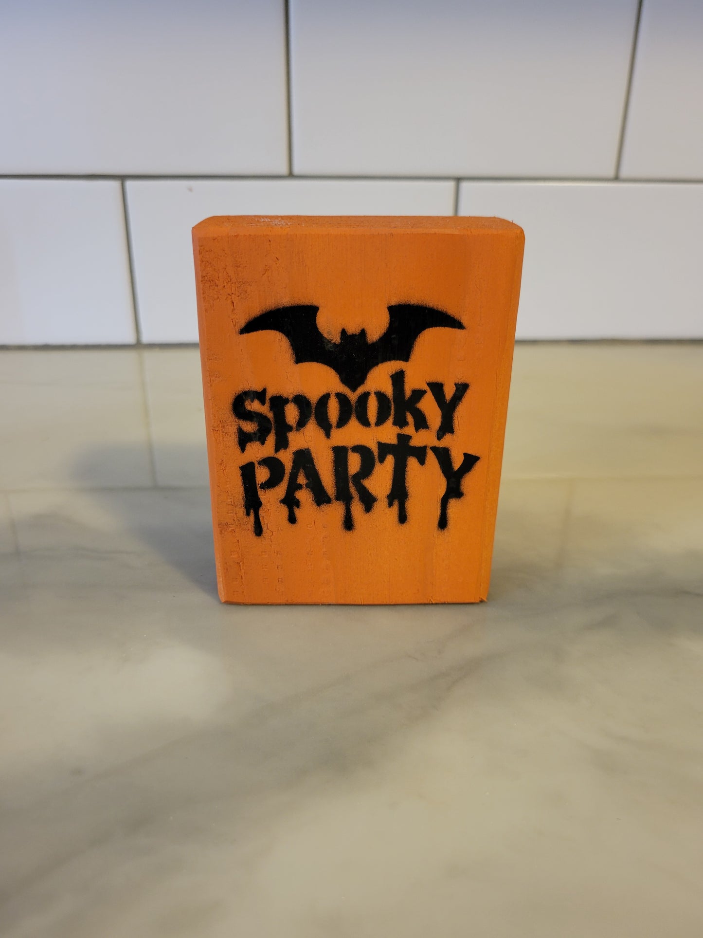 An orange rectangular wooden block sign with a black bat silhouette and the words 'Spooky Party' in a dripping gothic font, standing on a light marble surface against a white tiled wall.