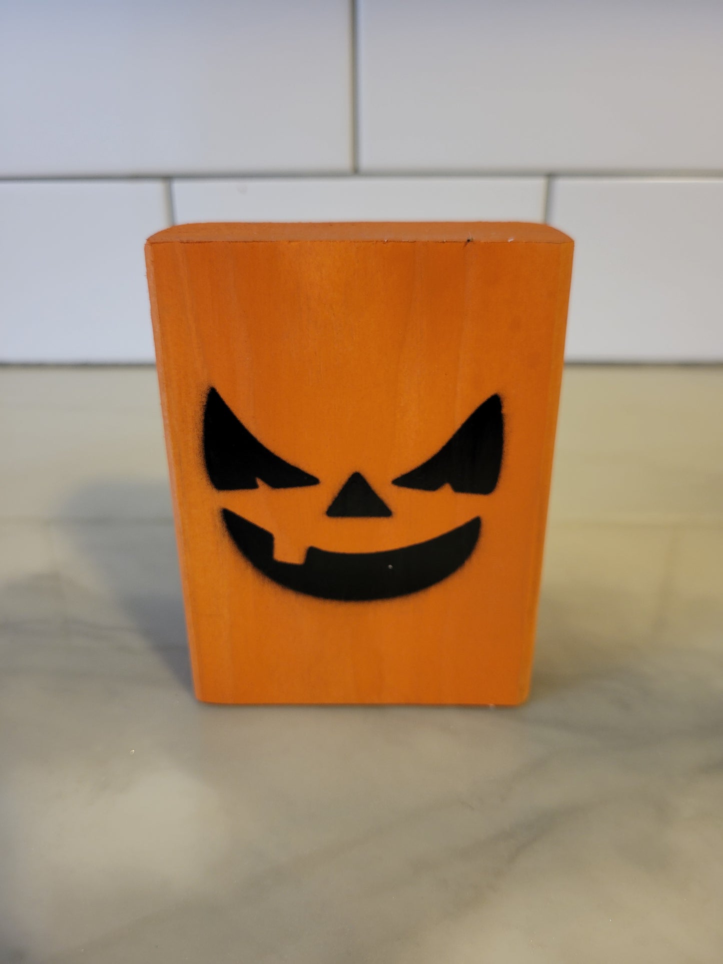 A small orange rectangular block with a black jack-o'-lantern face cutout, standing on a light marble-like surface in front of a white tiled wall.