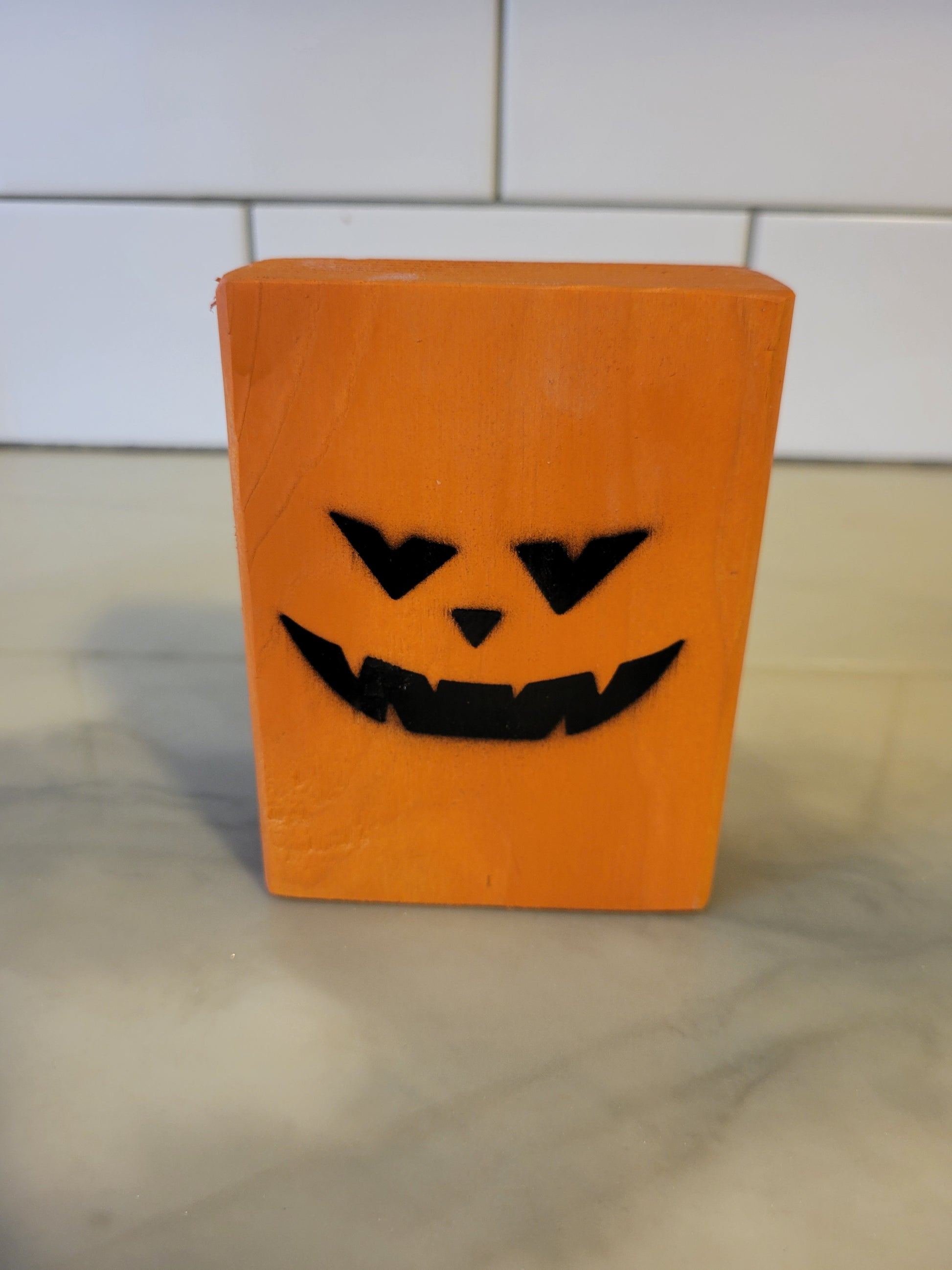 An orange rectangular block with a black jack-o'-lantern face cutout, standing on a light marble-like surface with a white tiled background.