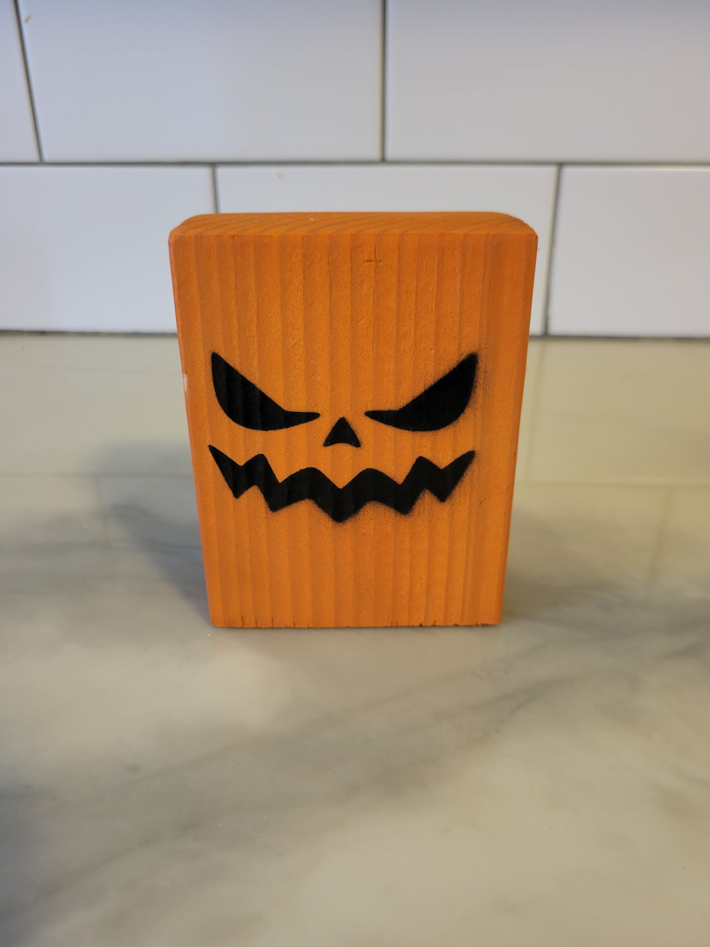 An orange rectangular block with a black carved jack-o'-lantern face, standing on a light marble-like surface with a white tiled wall in the background.