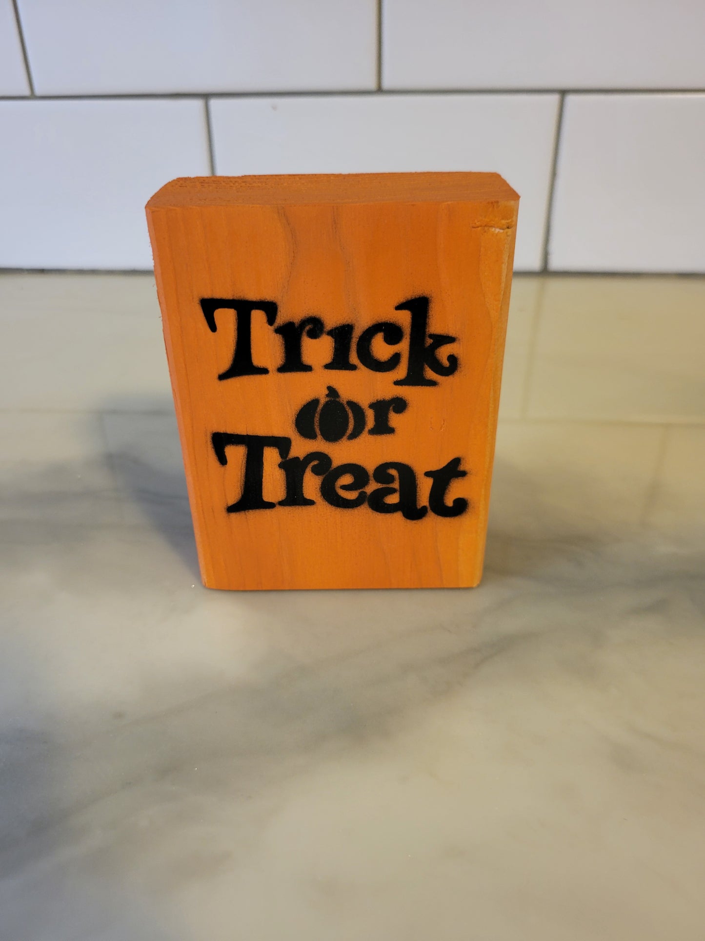 An orange rectangular wooden block sign with 'Trick or Treat' written in black stylized text, standing on a light marble-like surface with a white tiled background.