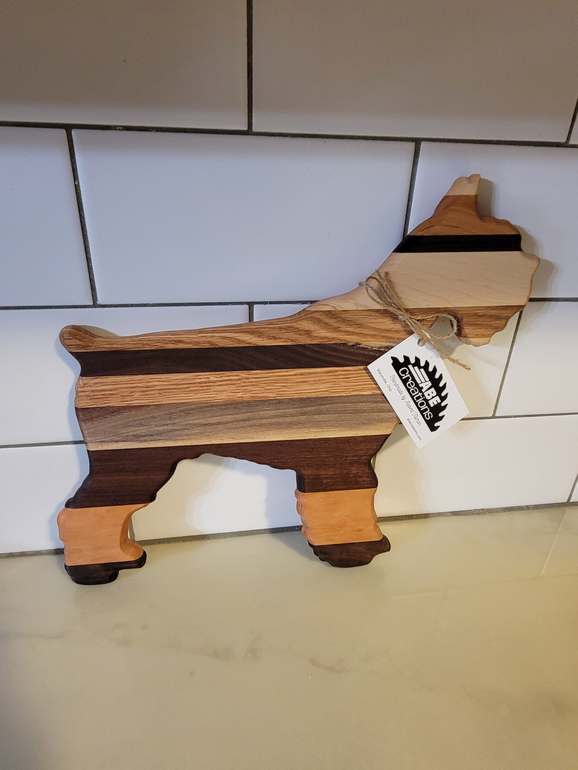 Wooden cutting board shaped like a dog on a tiled floor with a brand tag attached.
