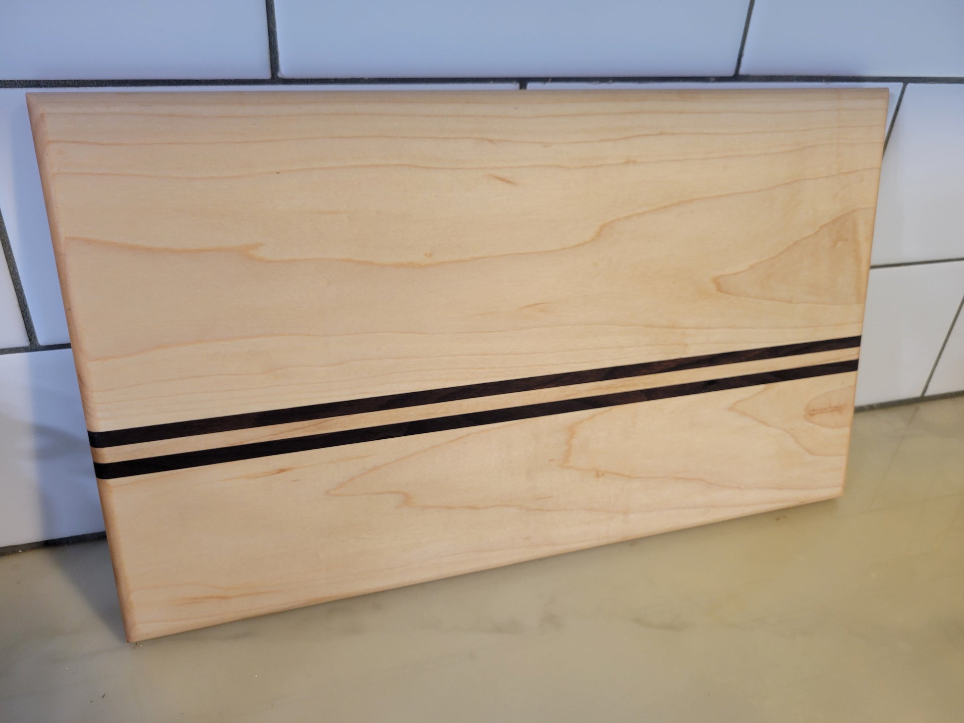 A rectangular light natural wood board with two parallel black stripes across the center, photographed against a tiled wall and light floor.