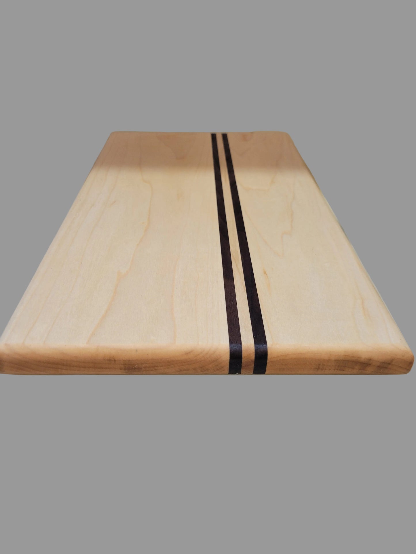 A rectangular wooden cutting board with a light natural maple finish and two dark brown stripes in the center, shown standing vertically against a tiled wall.