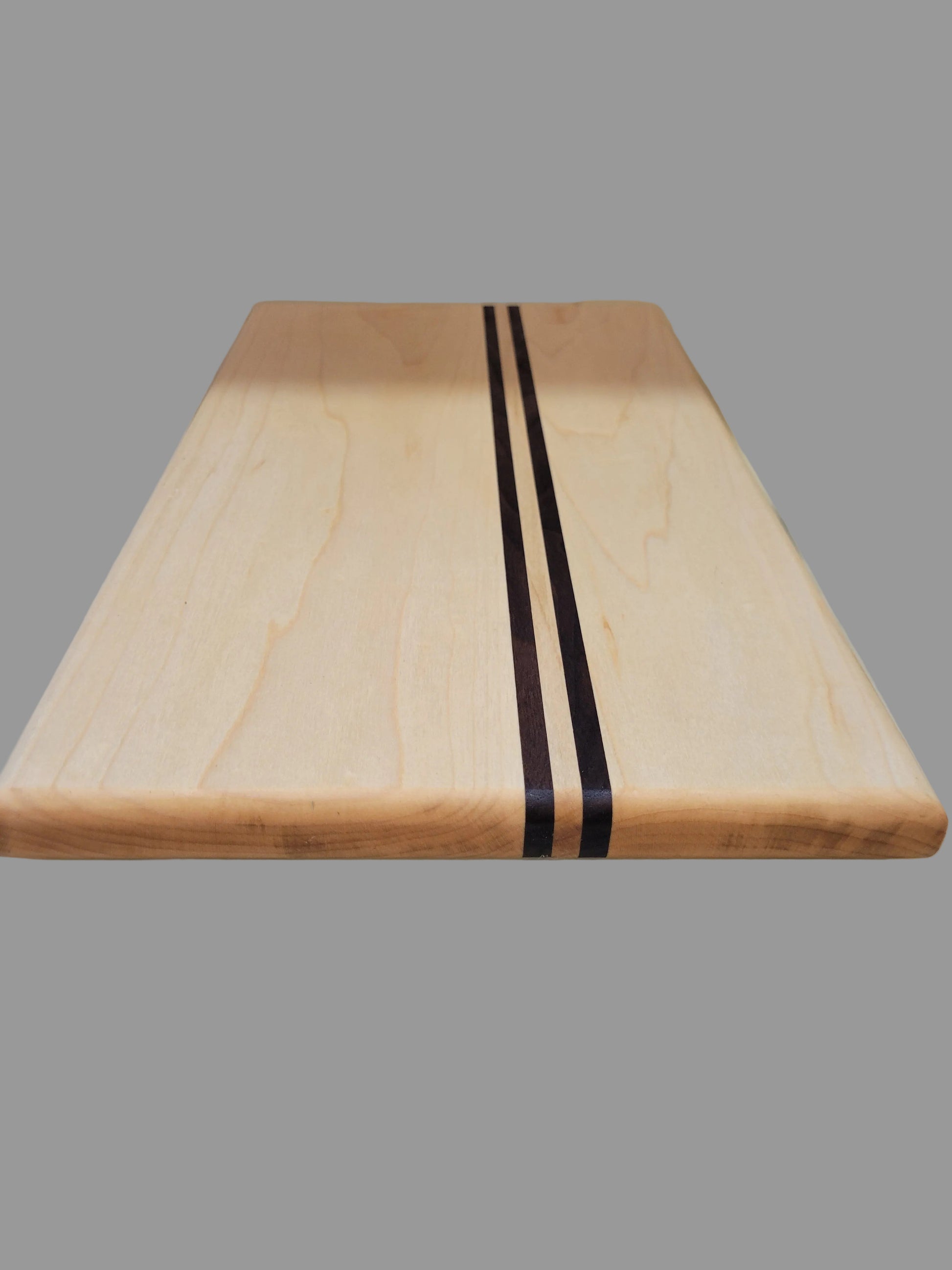 A rectangular wooden cutting board with a light natural maple finish and two dark brown stripes in the center, shown standing vertically against a tiled wall.