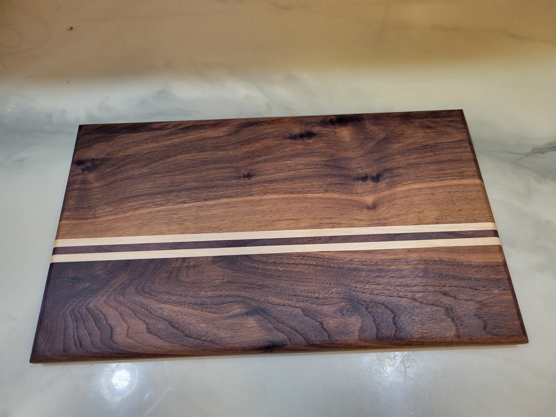 A rectangular wooden board with a dark brown walnut wood grain finish and two horizontal light beige inlay stripes near the center, photographed on a light marble-like surface.